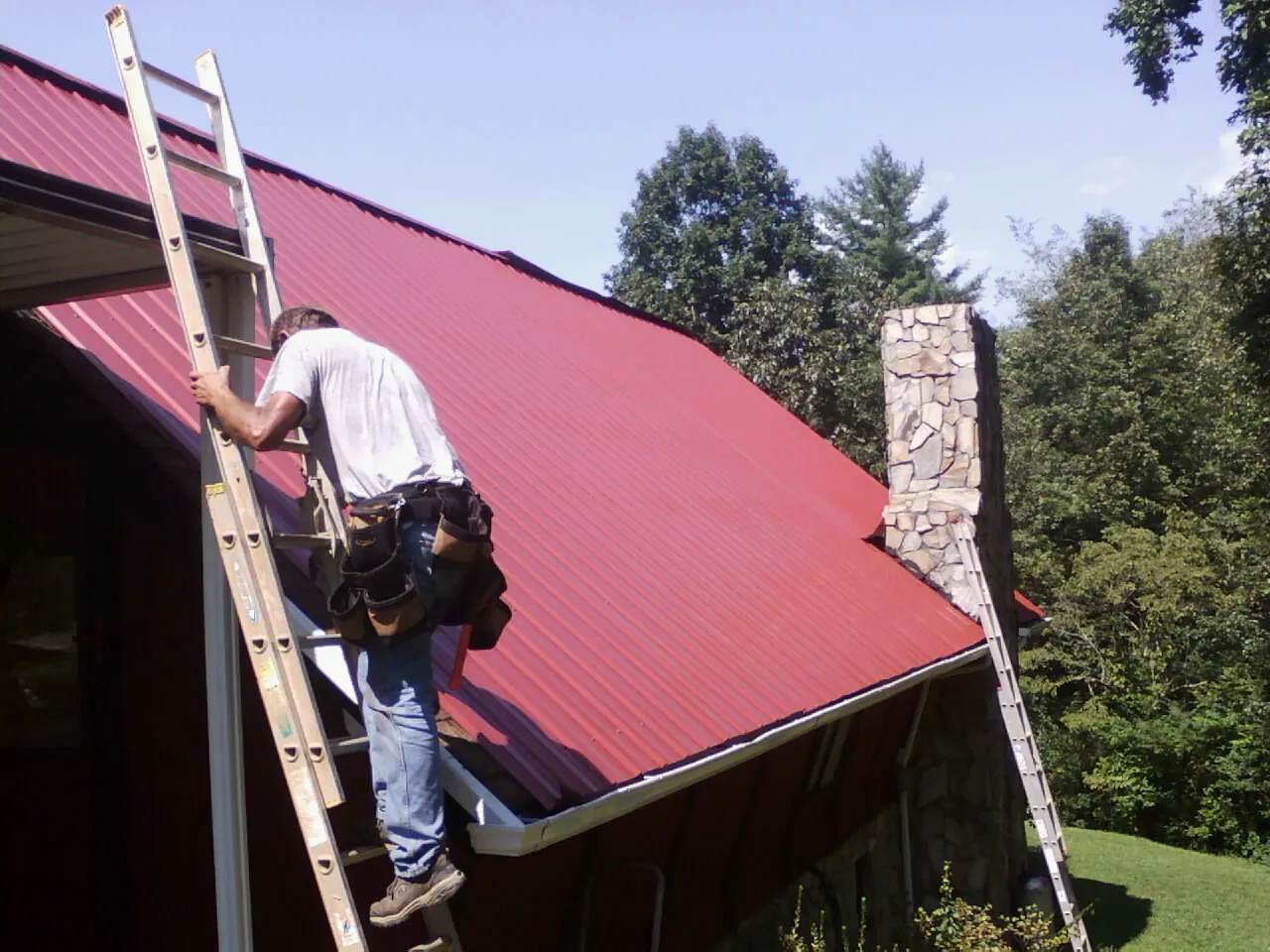 Roofer on a ladder beside a red metal roof and stone chimney — Soffit & Fascia Repair by Roofing Brothers in Osceola Mills